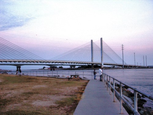 A cable-stayed bridge spans a river at sunset, with a walkway and people along the shore.