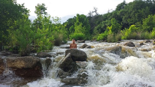 A child stands on a rock in a flowing river, surrounded by lush greenery and splashing water.
