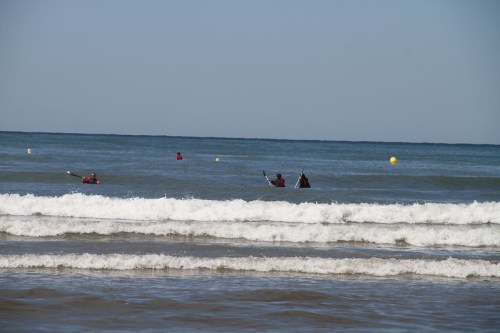 Two people in wetsuits are kayaking in the ocean, with gentle waves and a clear blue sky in the background.