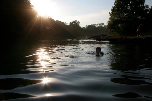 A child swims in a calm river at sunset, surrounded by trees and gentle ripples on the water's surface.