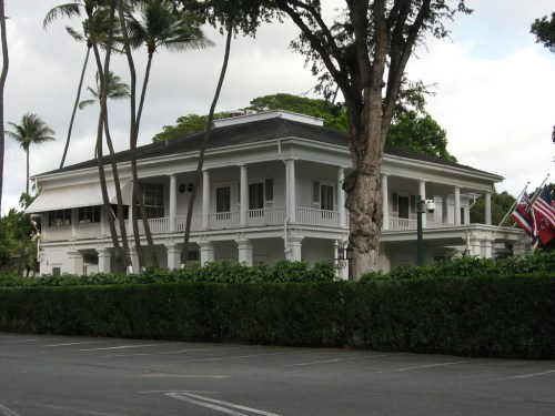A white colonial-style building surrounded by palm trees and greenery, with flags displayed in front.