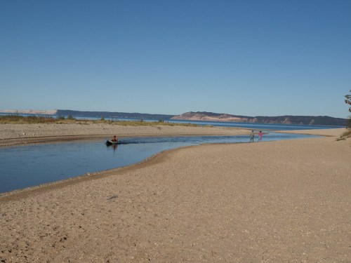 A serene river scene with a sandy shore, a small boat, and two people walking along the water's edge under a clear blue sky.