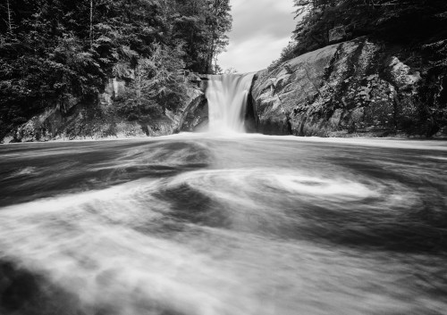 A serene black-and-white image of a waterfall cascading over rocks into a swirling pool below, surrounded by trees.