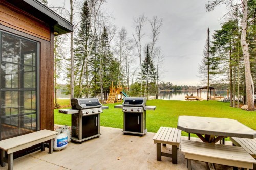 Two grills on a patio overlooking a serene lake, surrounded by trees and a grassy area with picnic tables.