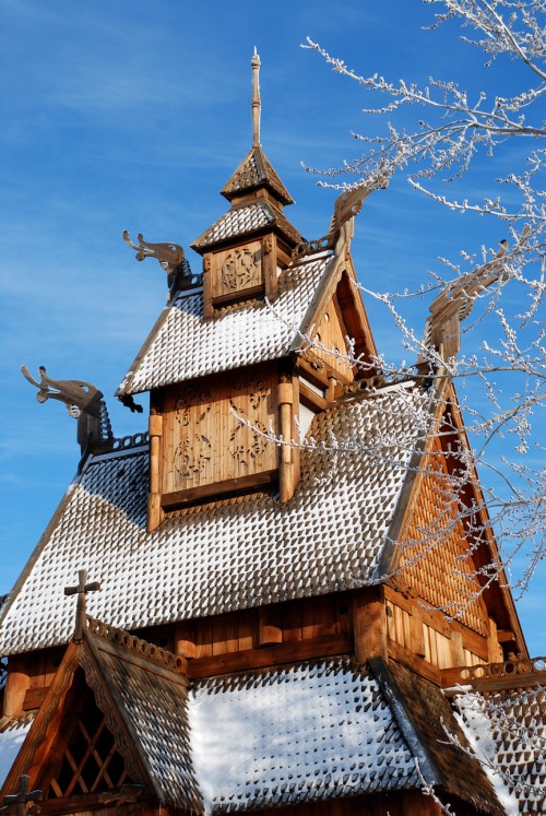 A wooden church with a steep, snow-covered roof and ornate carvings, set against a clear blue sky.