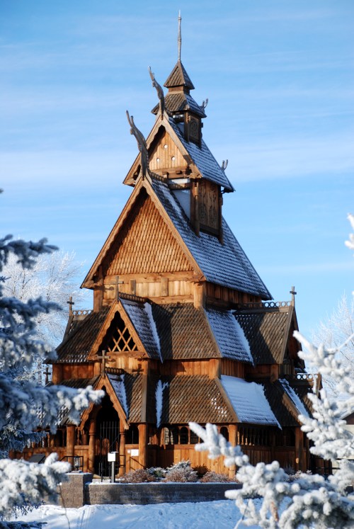A wooden, intricately designed building with a steep roof, surrounded by snow-covered trees under a clear blue sky.