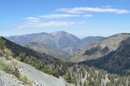 A panoramic view of rugged mountains and valleys under a clear blue sky, with scattered trees in the foreground.