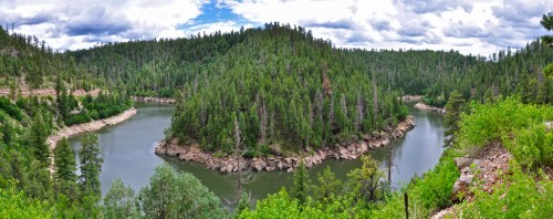 A panoramic view of a winding river surrounded by lush green forests and hills under a cloudy sky.