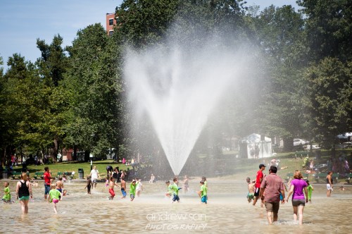 Children and families play in a splash pad, enjoying water sprays on a sunny day in a park.
