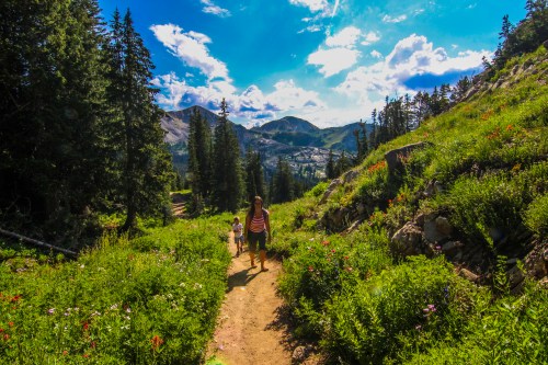 A woman and child walk along a dirt path through a vibrant green landscape with mountains and blue skies in the background.