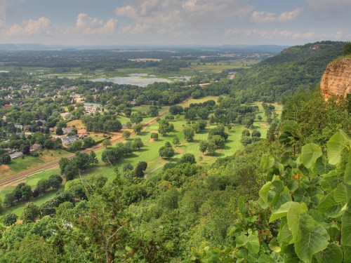 A panoramic view of a lush green landscape with a golf course, hills, and a river in the distance under a cloudy sky.