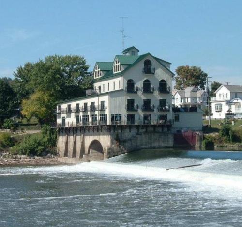 A riverside building with a green roof, balconies, and a waterfall in front, surrounded by trees and houses.