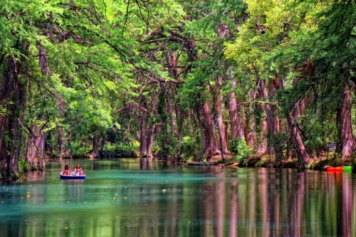 Two people in a small boat on a serene, tree-lined river with vibrant green foliage reflecting in the water.