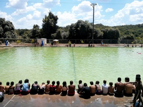A group of people sits by a green swimming pool under a sunny sky, surrounded by trees and hills.