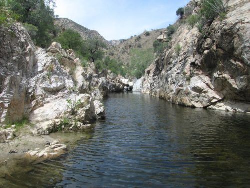 A serene river flows between rocky cliffs, surrounded by greenery under a clear blue sky.