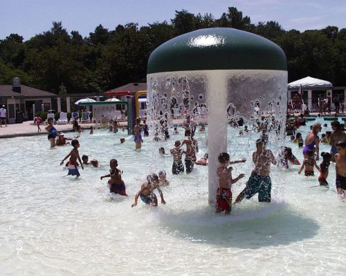 Children play in a splash pool under a large water feature, surrounded by trees and umbrellas on a sunny day.