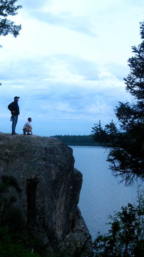 Two people stand on a rocky ledge overlooking a calm lake, surrounded by trees and a cloudy sky.