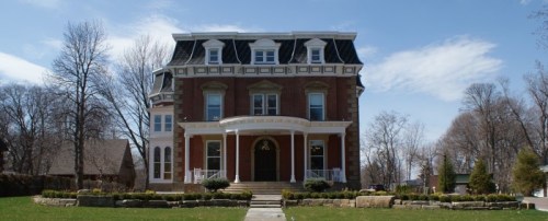 A large, historic brick house with a grand entrance, surrounded by trees and a well-maintained lawn under a clear blue sky.