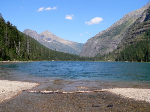 A serene lake surrounded by mountains and lush forests under a clear blue sky.