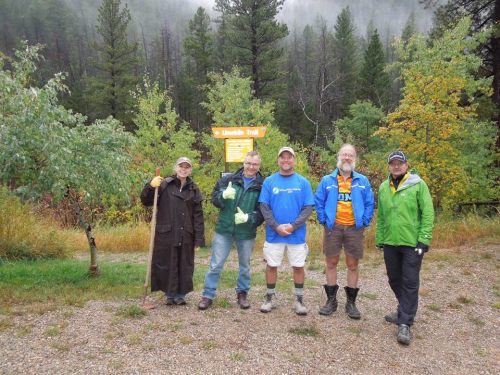 Five people stand at the start of a hiking trail, surrounded by trees and misty weather, smiling and posing for the photo.