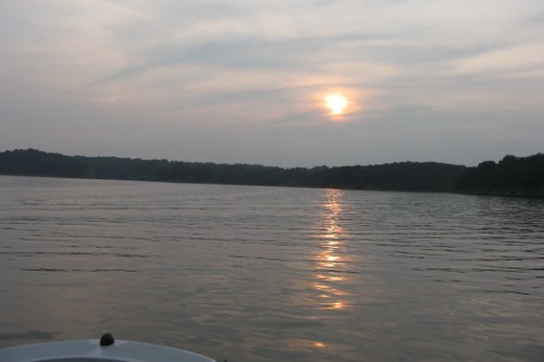 A serene lake at sunset, with soft reflections on the water and a hazy sky in the background.
