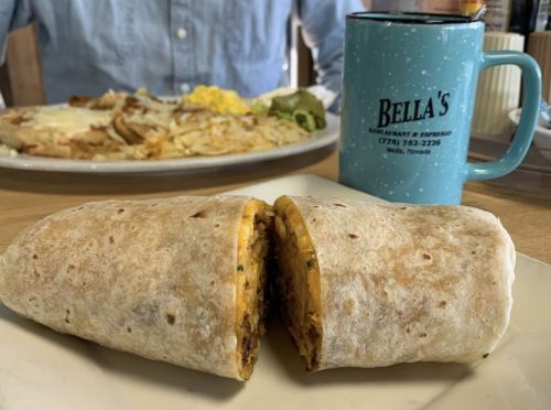 A cut burrito filled with eggs and meat, with a coffee mug and a plate of breakfast in the background.