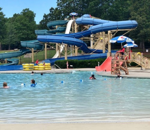 A water park scene featuring slides, a pool with swimmers, and colorful umbrellas in a green park setting.