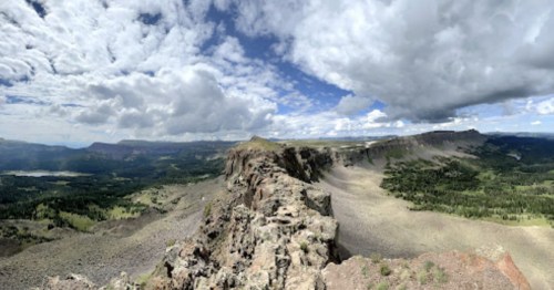 A panoramic view of a rugged mountain landscape under a partly cloudy sky, showcasing green valleys and rocky terrain.