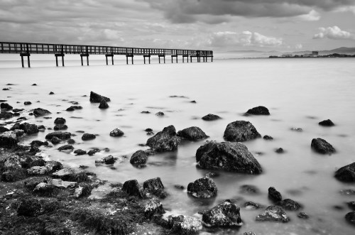 Black and white image of a calm shoreline with a wooden pier extending over smooth water and rocky foreground.