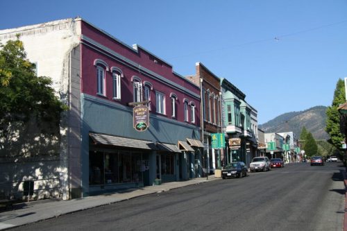 A quiet street lined with colorful historic buildings and shops, with mountains in the background under a clear blue sky.