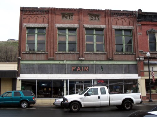 Historic brick building with large windows, featuring a sign that reads "FAIR" and a white truck parked in front.