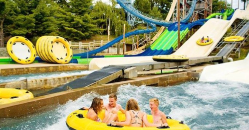 Four people enjoy a water ride in yellow tubes at a water park, with slides and greenery in the background.