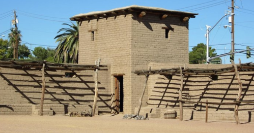 A rustic adobe building with a tower, surrounded by wooden beams and palm trees under a clear blue sky.