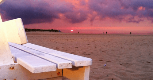 A beach scene at sunset, featuring a white bench and vibrant pink and purple skies with silhouettes of people in the distance.