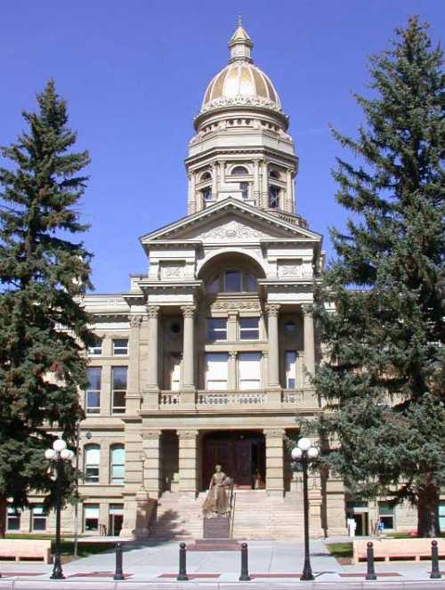 Historic building with a golden dome, flanked by tall trees and a statue at the entrance, under a clear blue sky.