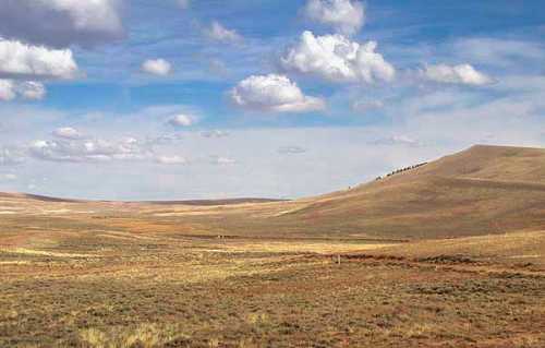 A vast, open landscape with rolling hills, grassy plains, and a blue sky dotted with fluffy white clouds.