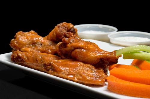 A plate of saucy chicken wings with carrot sticks and two dipping sauces on the side, set against a black background.