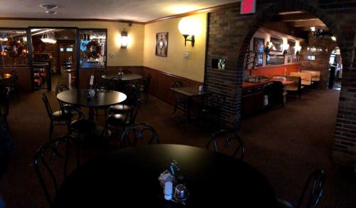 Interior of a cozy restaurant featuring black tables, chairs, and warm lighting, with a brick wall and a dining area.