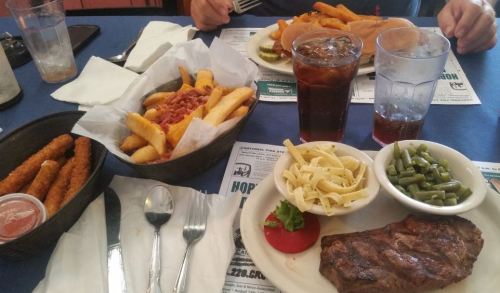 A table with a variety of dishes: steak, pasta, green beans, fries, and drinks, set in a casual dining environment.