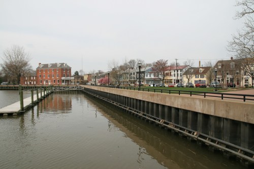 A calm waterfront scene featuring a dock, trees, and historic buildings along the shore under a cloudy sky.