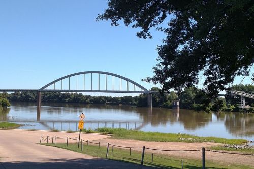 A bridge arches over a calm river, surrounded by greenery and a clear blue sky.