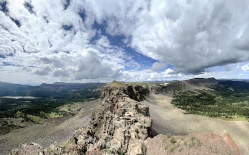 A panoramic view of a rocky landscape under a cloudy sky, with mountains and greenery in the distance.