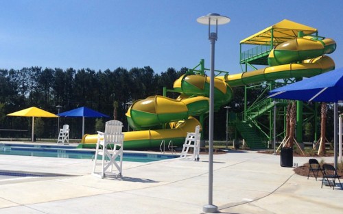 A bright outdoor pool area featuring a large yellow and green water slide, lounge chairs, and blue umbrellas.