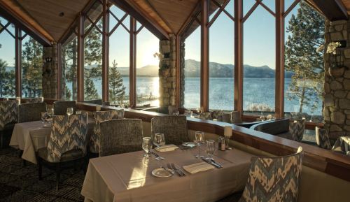 Elegant restaurant interior with tables set for dining, large windows showcasing a scenic lake and mountains at sunset.