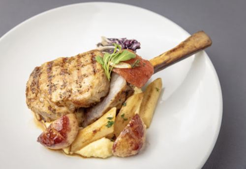 Grilled pork chop with a bone, served with blood orange, potatoes, and greens on a white plate.