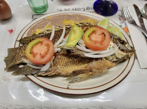 A plate of fried fish garnished with sliced tomatoes, onions, and lime wedges, served on a decorative tablecloth.
