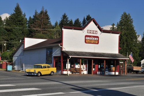 A vintage grocery store with a yellow car parked outside, surrounded by trees and a clear blue sky.