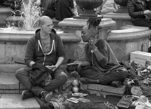 A man and a woman sit on the ground near a fountain, engaged in conversation, surrounded by various items.