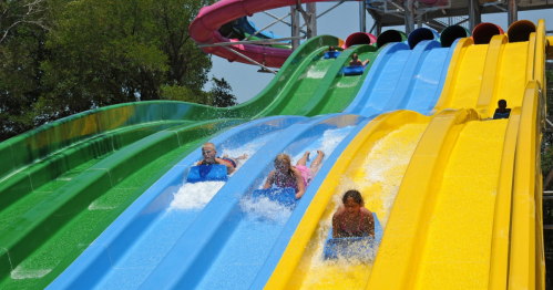 Four people slide down colorful water slides, splashing into a pool below on a sunny day.