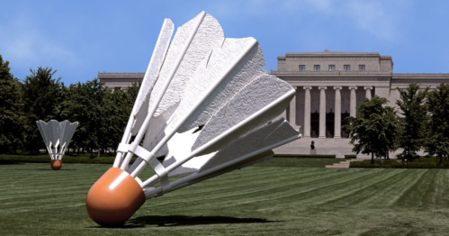 Two large shuttlecock sculptures on a grassy lawn in front of a classical building under a clear blue sky.